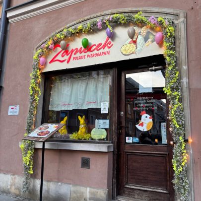Colorful storefront of a local zapiekanka restaurant decorated with seasonal flowers and inviting street-side charm.