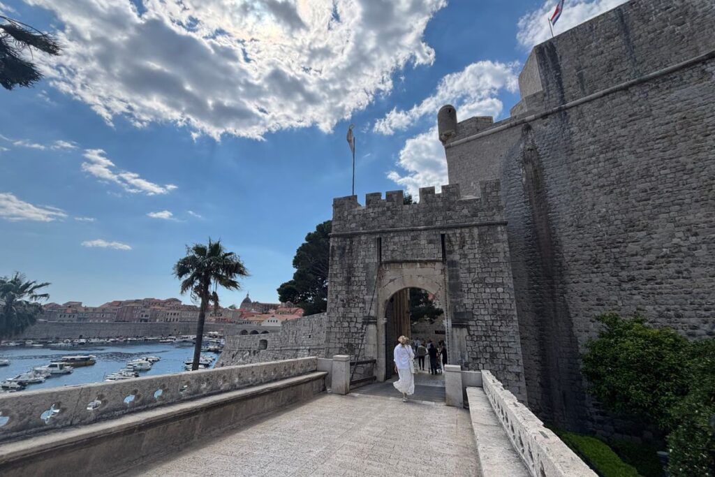 Entrance to Dubrovnik Old Town through the historic Pile Gate, with medieval city walls, palm trees, and views of Croatia's Adriatic coast.