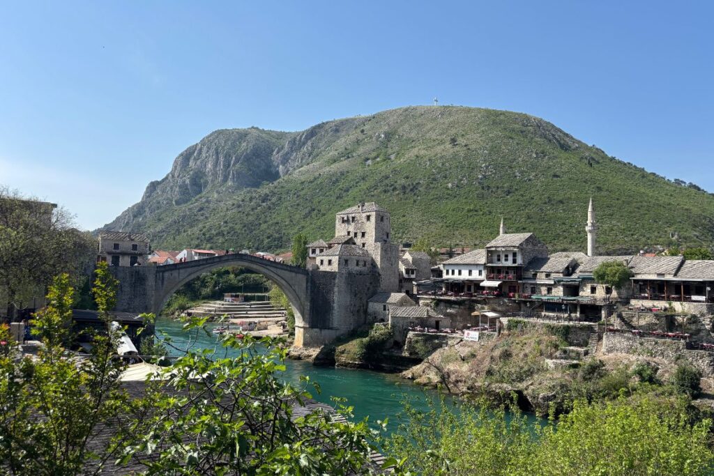 Stari Most bridge spanning the turquoise Neretva River in Mostar, Bosnia and Herzegovina, with stone buildings and green mountains in the background.