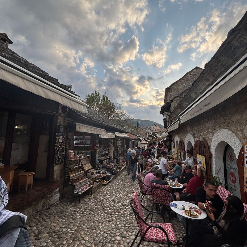 Outdoor cafés and shops lining a cobblestone street in Mostar's Old Town at sunset.