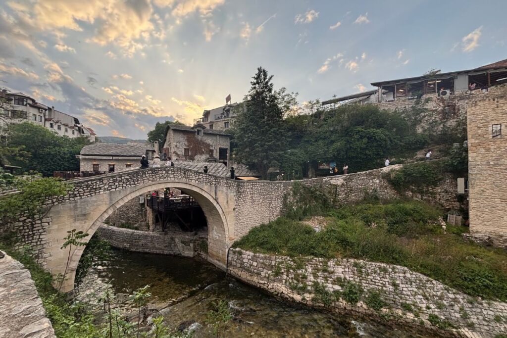 Kriva Ćuprija (Crooked Bridge) in Mostar's Old Town, a small stone arch bridge crossing a narrow stream beneath a colorful sunset sky.