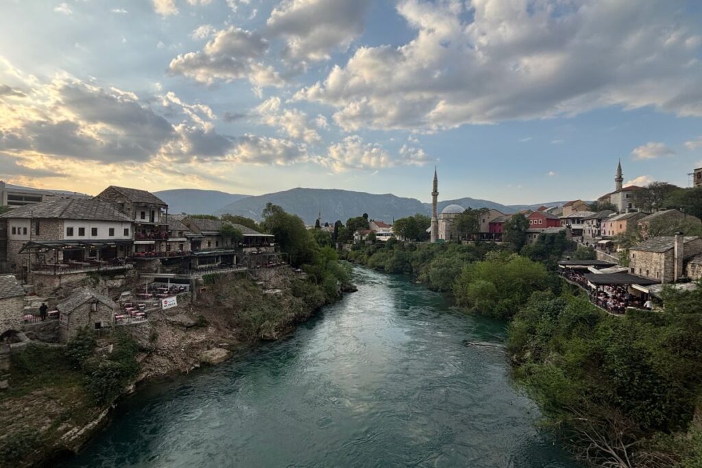 Evening view along the Neretva River in Mostar, Bosnia and Herzegovina, with historic stone buildings, a mosque minaret, and mountains beneath a dramatic sunset sky.