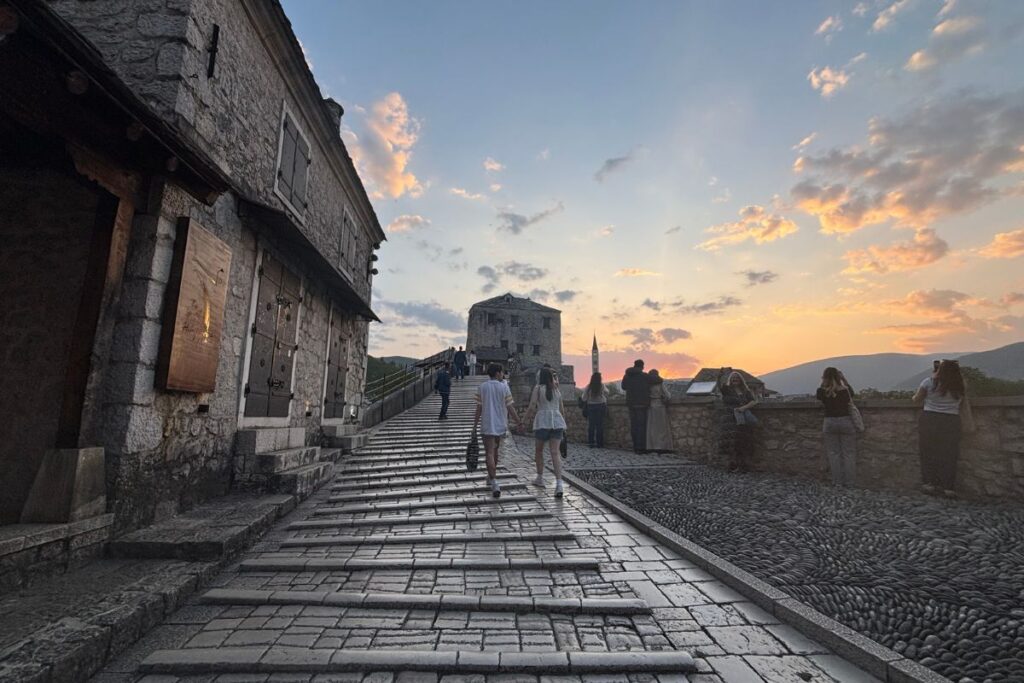 Sunset view along the stone walkway of Mostar's Old Bridge with visitors overlooking the Neretva River in Bosnia and Herzegovina.