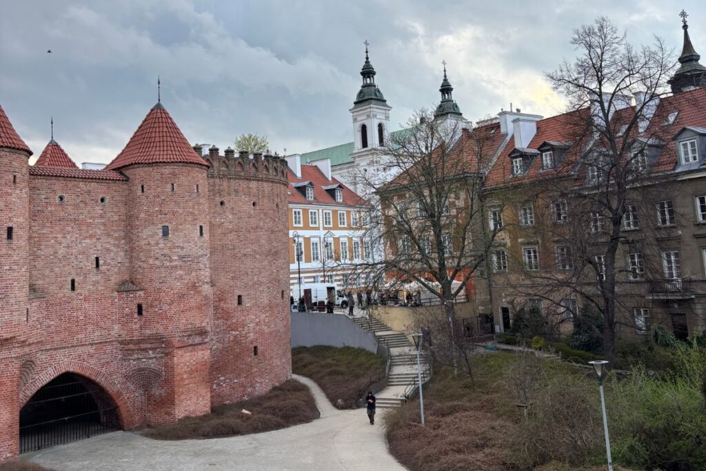 Historic red brick Barbican walls and towers in Warsaw Old Town, surrounded by colorful townhouses and church spires beneath a dramatic spring sky.