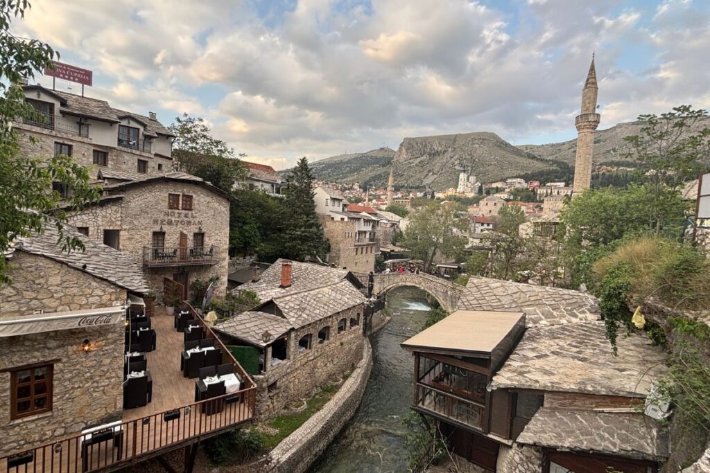 Historic stone rooftops and winding waterways in Mostar's Old Town, with the Crooked Bridge, mosque minaret, and surrounding mountains glowing beneath an evening sky.