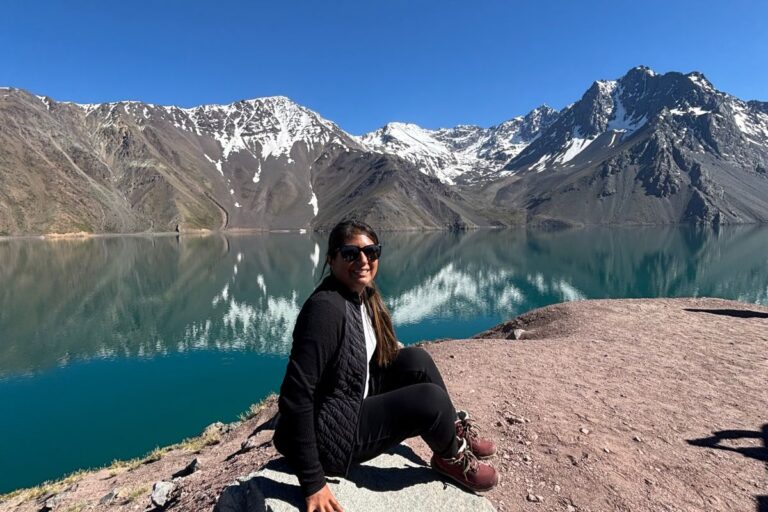 Traveler sitting beside the turquoise waters of Embalse El Yeso in the Chilean Andes with snow-capped mountains reflected in the lake.