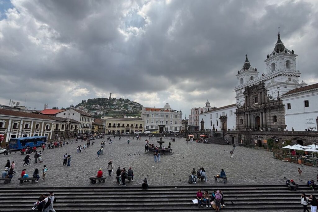 View of Plaza de la Independencia in Quito, Ecuador, with the Metropolitan Cathedral, colonial buildings, and El Panecillo overlooking the historic center beneath dramatic clouds.