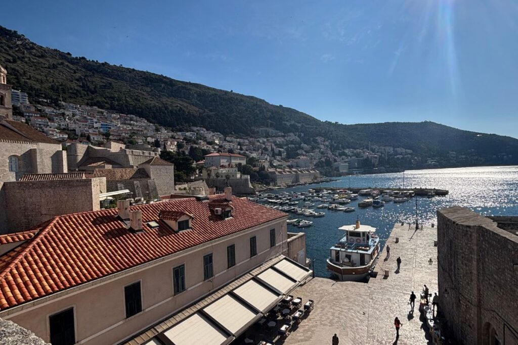 View over Dubrovnik's Old Town harbor, with terracotta rooftops, stone walls, fishing boats, and the sparkling Adriatic Sea beneath a clear blue sky.