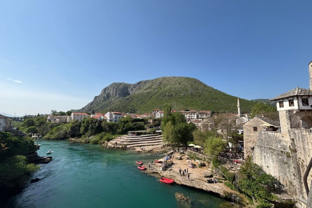 The turquoise Neretva River flowing through Mostar, Bosnia and Herzegovina, with historic stone buildings, a mosque minaret, and green mountains rising above the old town.