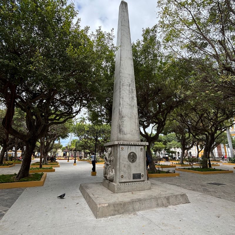 Stone obelisk surrounded by mature shade trees in Granada's Central Park on a peaceful afternoon.