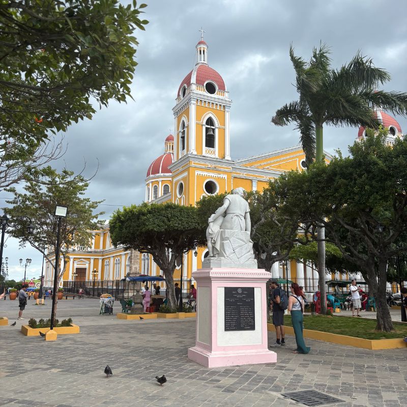 Statue in the foreground with Granada Cathedral rising behind it, overlooking the city's main plaza under dramatic clouds.