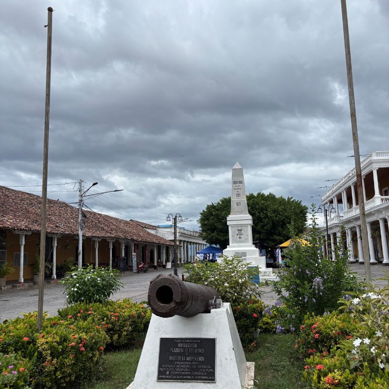 Historic cannon and monument in Granada's Central Park, framed by colorful colonial buildings beneath a cloudy sky.