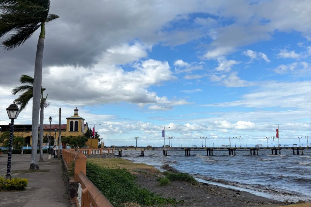 Palm-lined waterfront promenade along Lake Nicaragua in Granada with a pier, colorful colonial buildings, and dramatic clouds overhead.