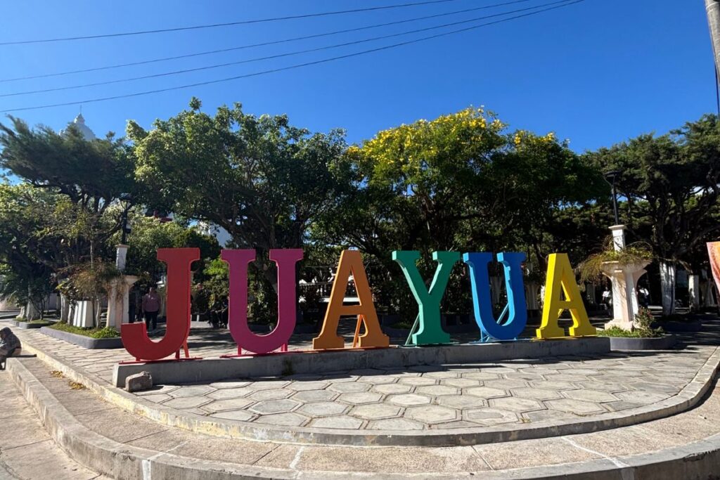 Colorful "Juayúa" town sign in the central plaza of Juayúa, El Salvador, surrounded by trees on a sunny day.