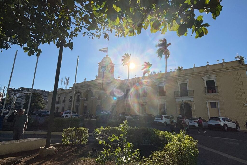 Sunlight shining over the historic municipal building and central plaza in Santa Ana, El Salvador, framed by palm trees and leafy branches.