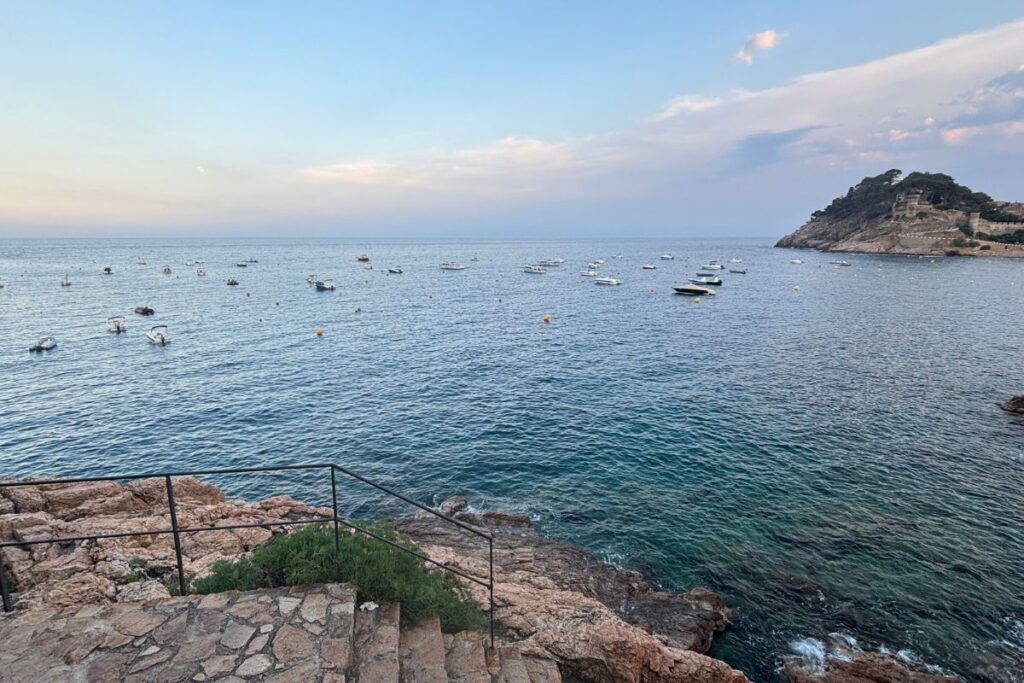 View of the Mediterranean Sea at sunset from the rocky coastline of Tossa de Mar on Spain's Costa Brava, with boats anchored offshore.