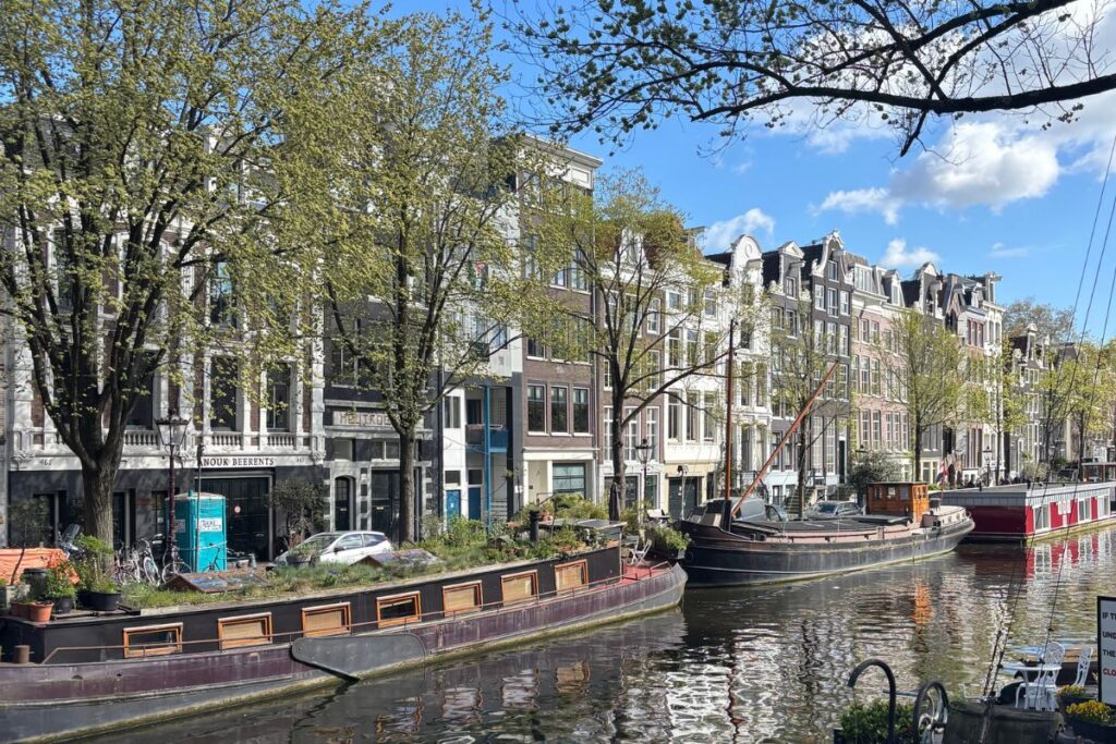 Canal lined with historic Amsterdam houses, houseboats, bicycles, and leafy trees on a sunny day.
