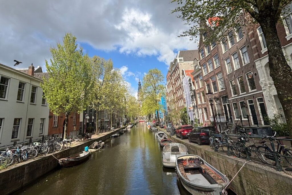 Narrow Amsterdam canal lined with historic houses, bicycles, trees, and small boats.