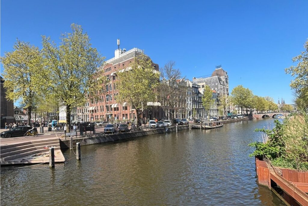 Wide Amsterdam canal surrounded by leafy trees and historic buildings under a bright blue sky.