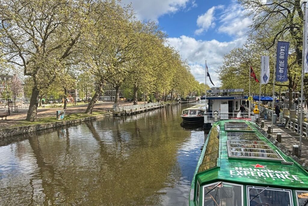 Tree-lined Amsterdam canal with canal cruise boats docked along the water.