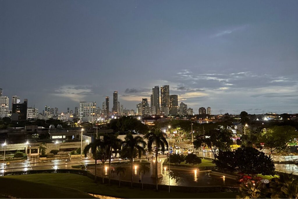 View of Cartagena's skyline at dusk with illuminated high-rise buildings, palm trees, and the historic city walls in the foreground after a rain shower.