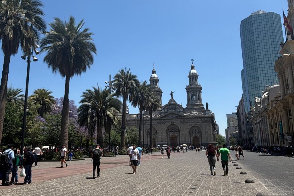 View of Plaza de Armas in Santiago, Chile, with palm trees, Santiago Metropolitan Cathedral, and the city's modern skyline in the background.