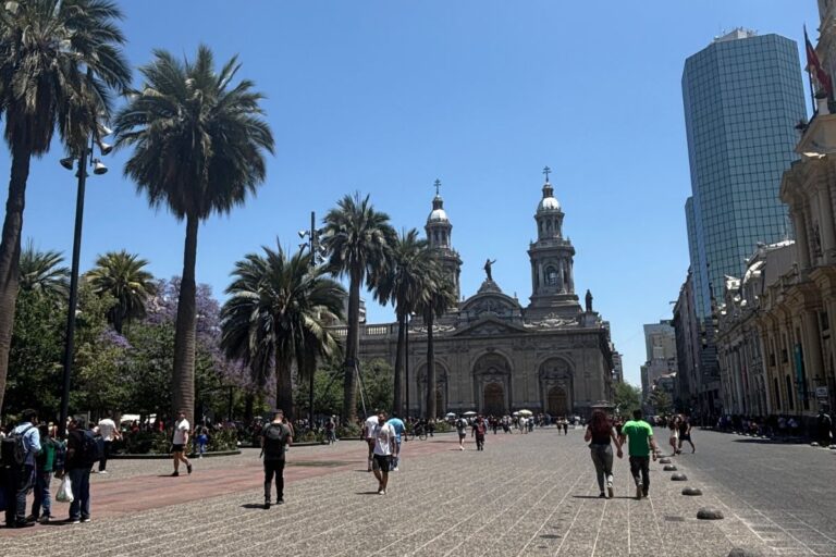 View of Plaza de Armas in Santiago, Chile, with palm trees, Santiago Metropolitan Cathedral, and the city's modern skyline in the background.