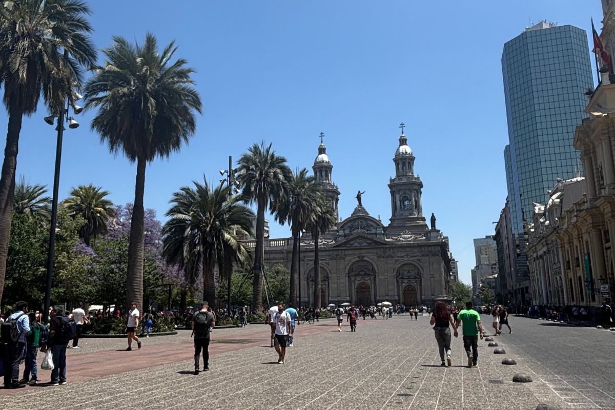 View of Plaza de Armas in Santiago, Chile, with palm trees, Santiago Metropolitan Cathedral, and the city's modern skyline in the background.