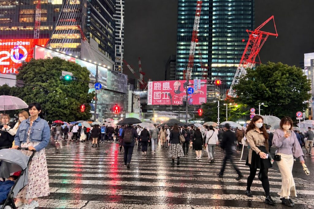 Shibuya Crossing at night in Tokyo, with crowds crossing the rain-covered intersection beneath illuminated buildings.